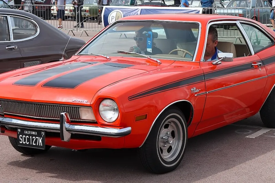 Red 1972 Ford Pinto with black racing stripes on display with man sitting in driver's seat.